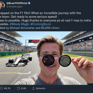 Brad Pitt takes a selfie on a Formula 1 track, smiling and holding up round sunglasses, with a white and orange F1 car and grandstands visible behind him.