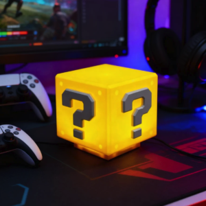 Glowing Super Mario Question Block lamp on a gaming desk with PS5 controllers.