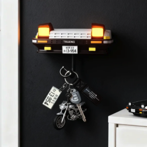 Toyota AE86 Trueno car front key holder with illuminated pop-up headlights, holding keys, next to a Mazda RX-7 model car.