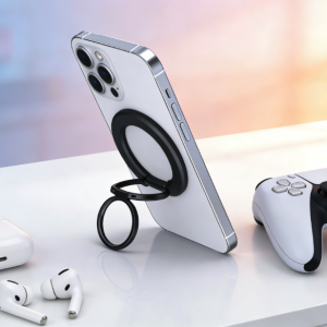 White iPhone standing with a black magnetic ring kickstand, alongside white AirPods and a PS5 controller on a clean white surface.