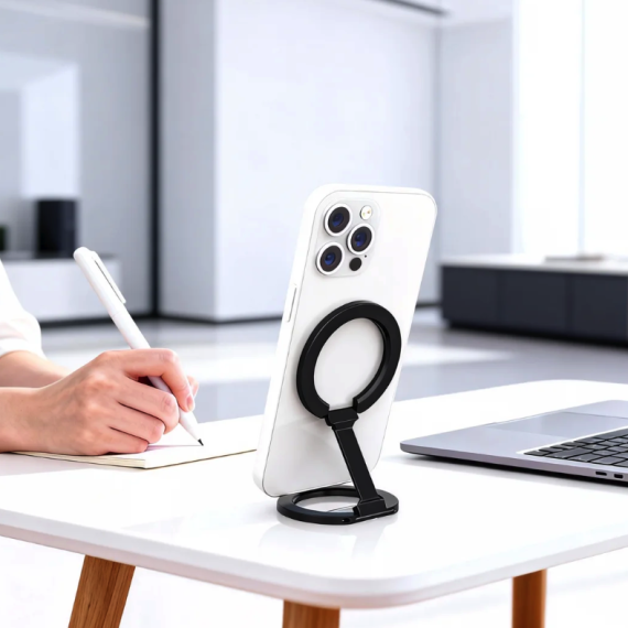 White iPhone with black ring stand on a desk, next to a person writing and a silver laptop, in a bright office.