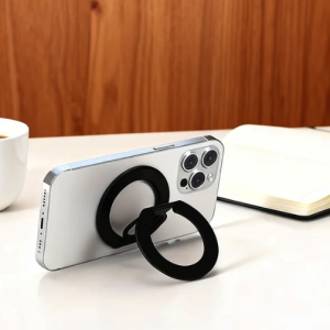 Silver smartphone with black magnetic ring kickstand, coffee mug, and open notebook on a white desk.