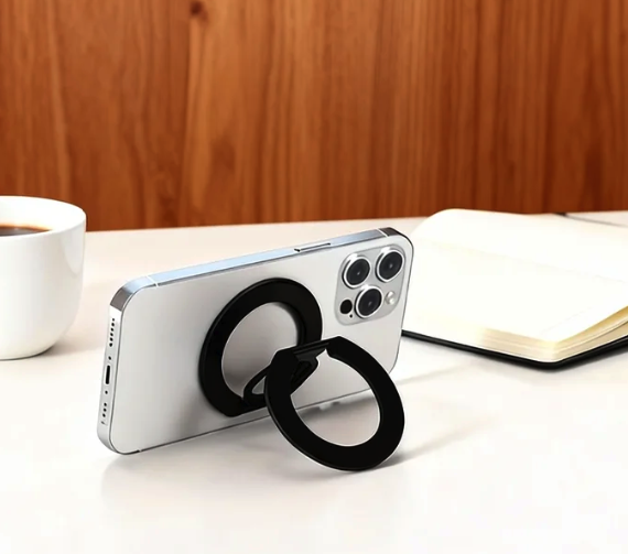 Silver smartphone with black magnetic ring kickstand, coffee mug, and open notebook on a white desk.
